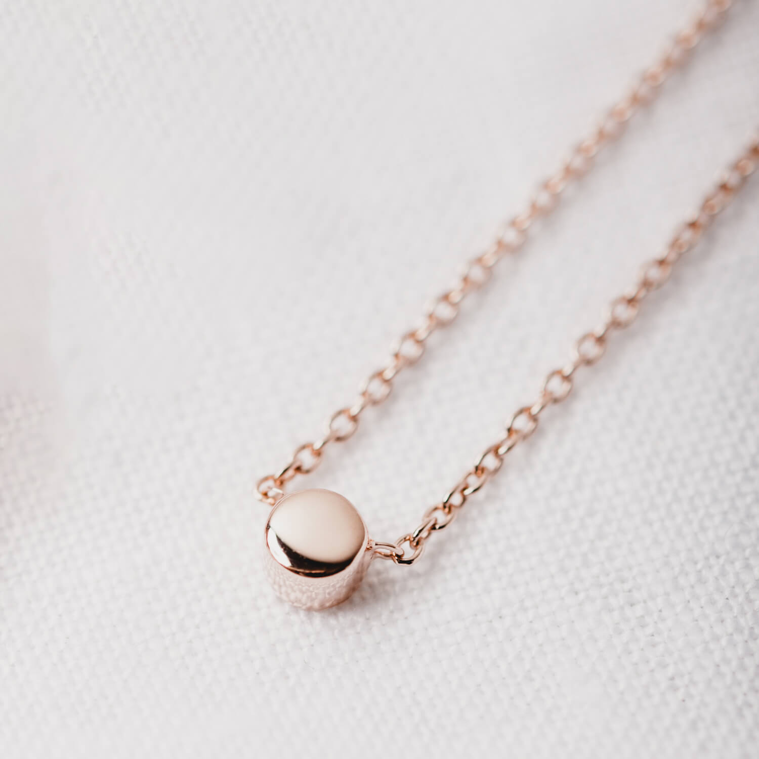 Close up of a rose gold matthew calvin dot necklace on a white textured background, the light reflecting off the pendant to show the sparkle