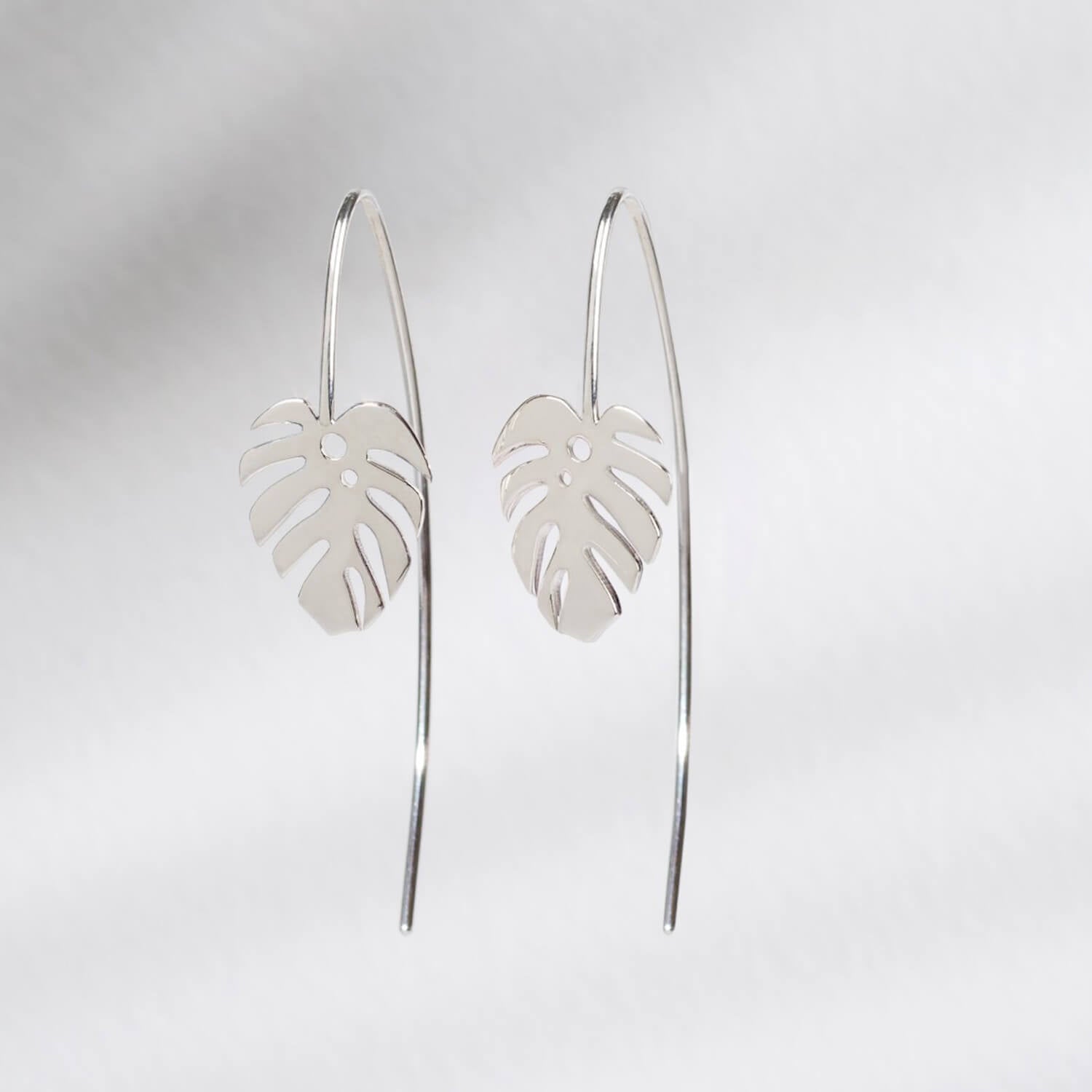 A close up of a pair of silver dropback earrings with large monstera leaf charms