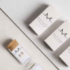 A pair of Matthew Calvin rose gold dot studs in a glass tube with cork stopper, alongside white branded carton packaging