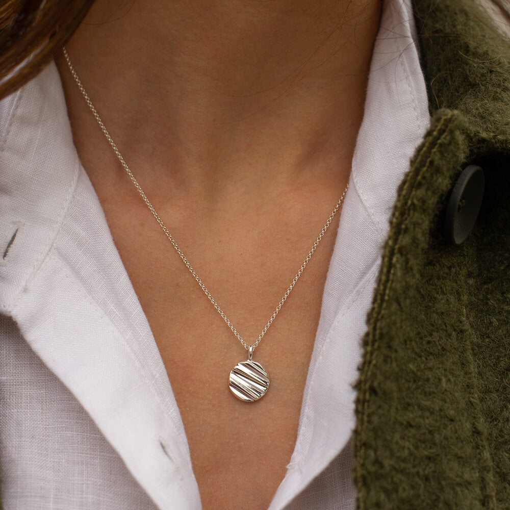 An Origins Necklace in silver being worn by a model in a green coat