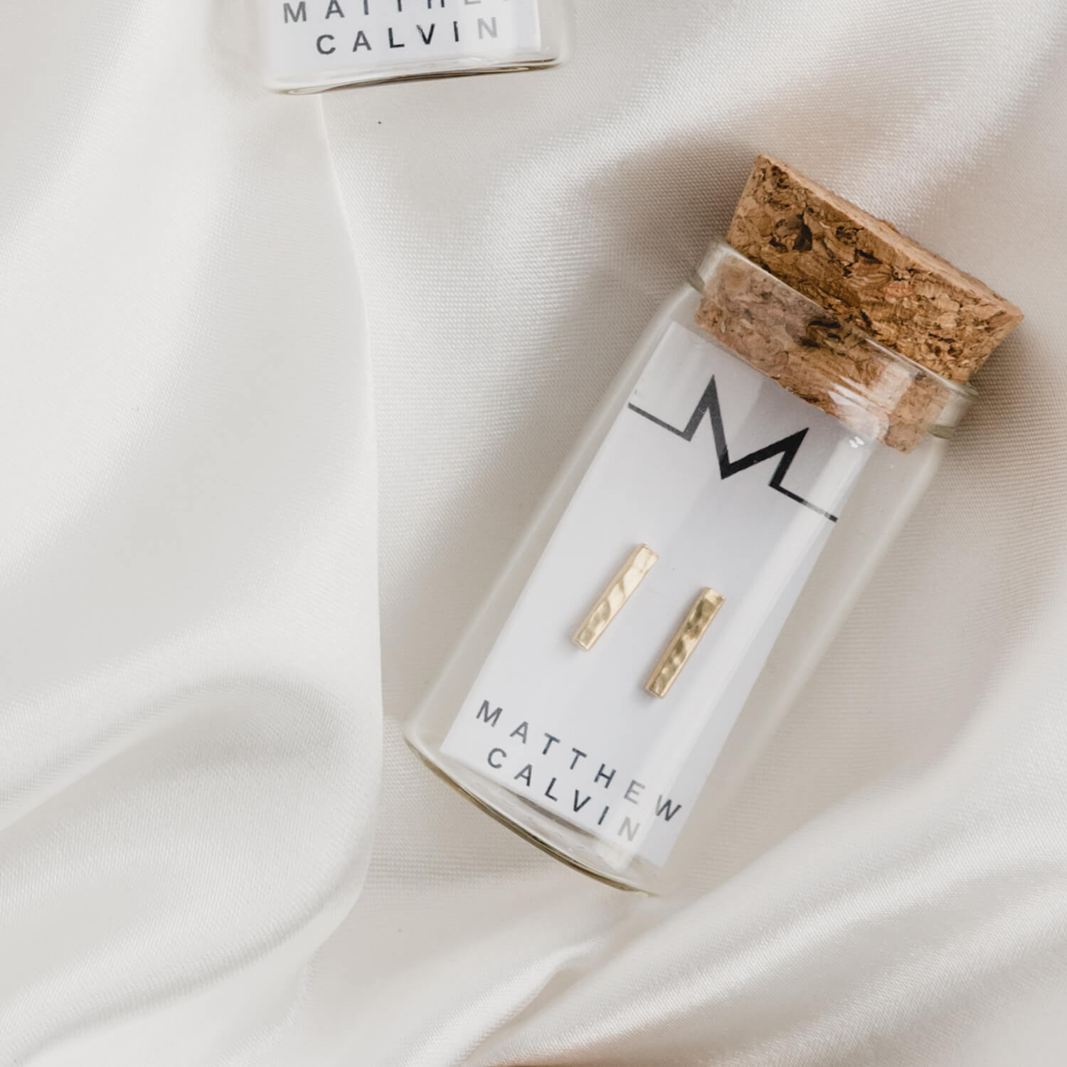 Small glass bottle with a cork lid, holding Textured Bar Earrings by Matthew Calvin in gold