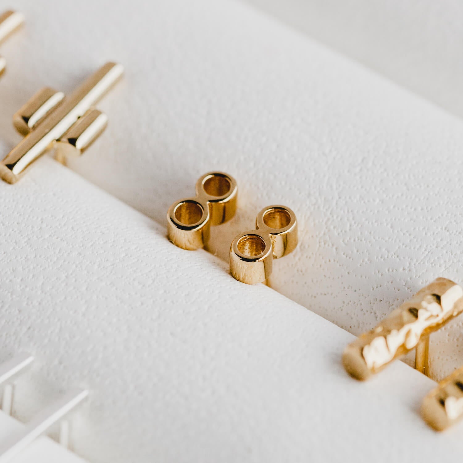 Close up of Gold tiny double tube stud earrings by matthew calvin, featuring other gold designs blurred out on the edges